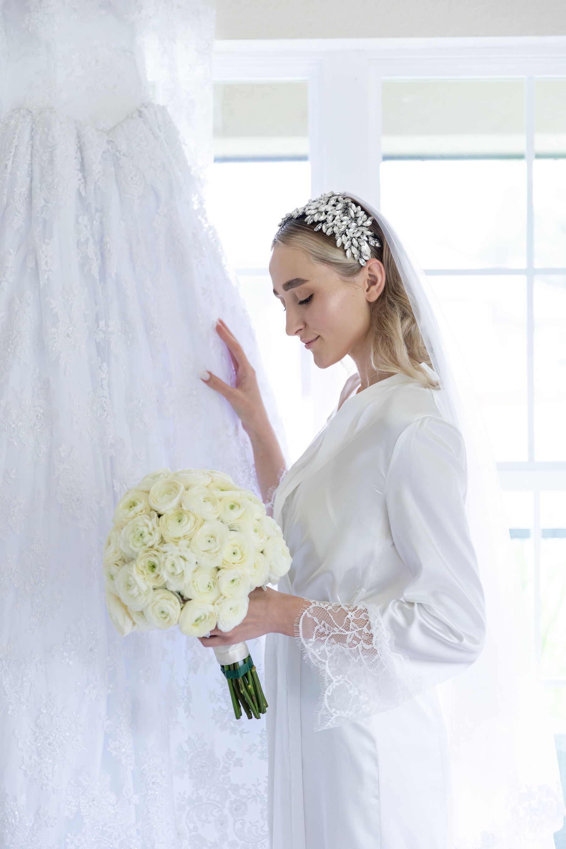 Bride with a jewelled headpiece holding white roses by a window