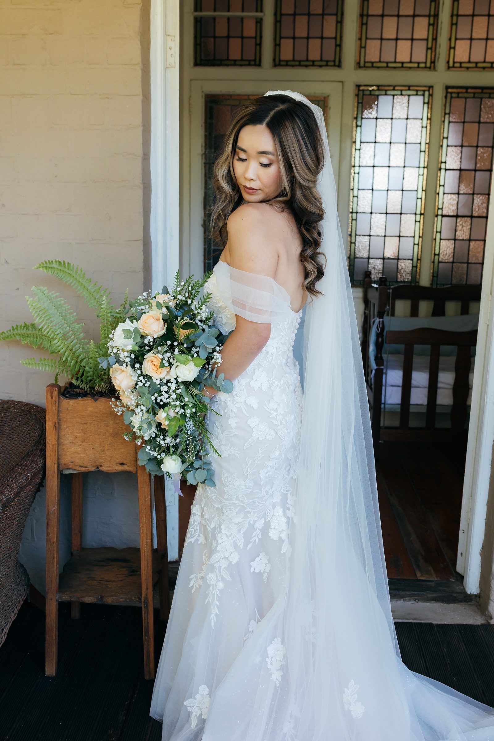 Bride in an off-shoulder gown holding her bouquet by a window